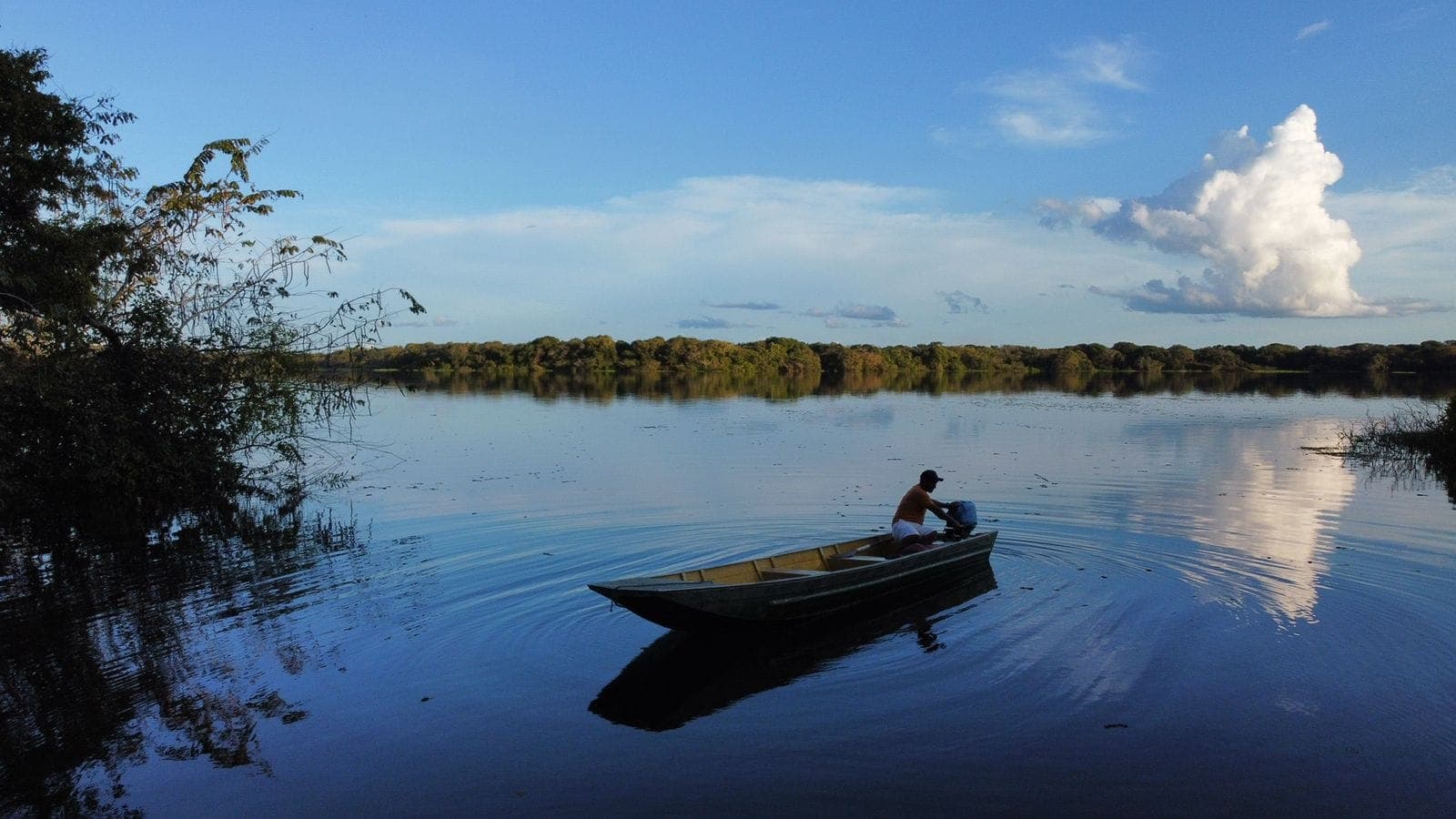 Homem numa canoa no rio, sob céu azul com nuvens, com a mata refletida na água.