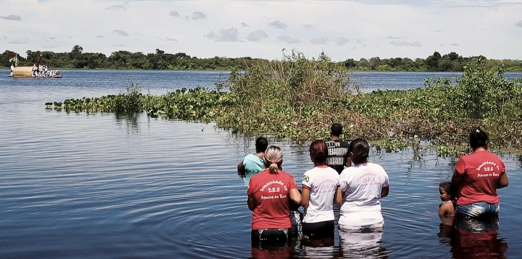 Pessoas de camisa vermelha e branca entrando no rio, de costas, perto de uma ilha de vegetação.