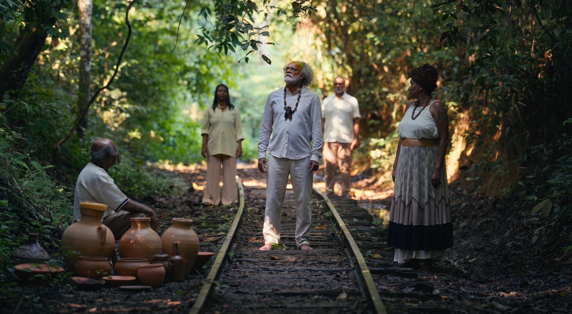Pessoas paradas sobre um trilho de trem no meio da mata, com potes de barro no chão.