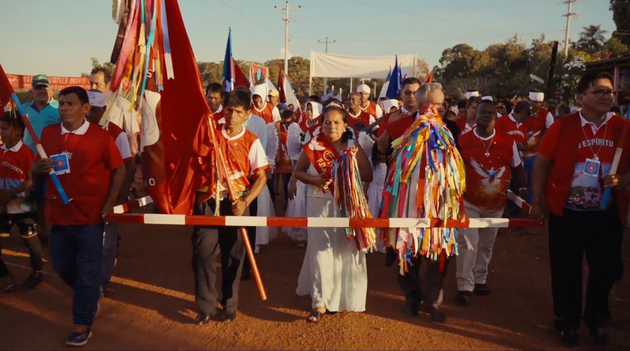 Procesión del Divino Espíritu Santo en una calle de tierra, con hombres y mujeres de camisa roja, banderas y cintas de colores.