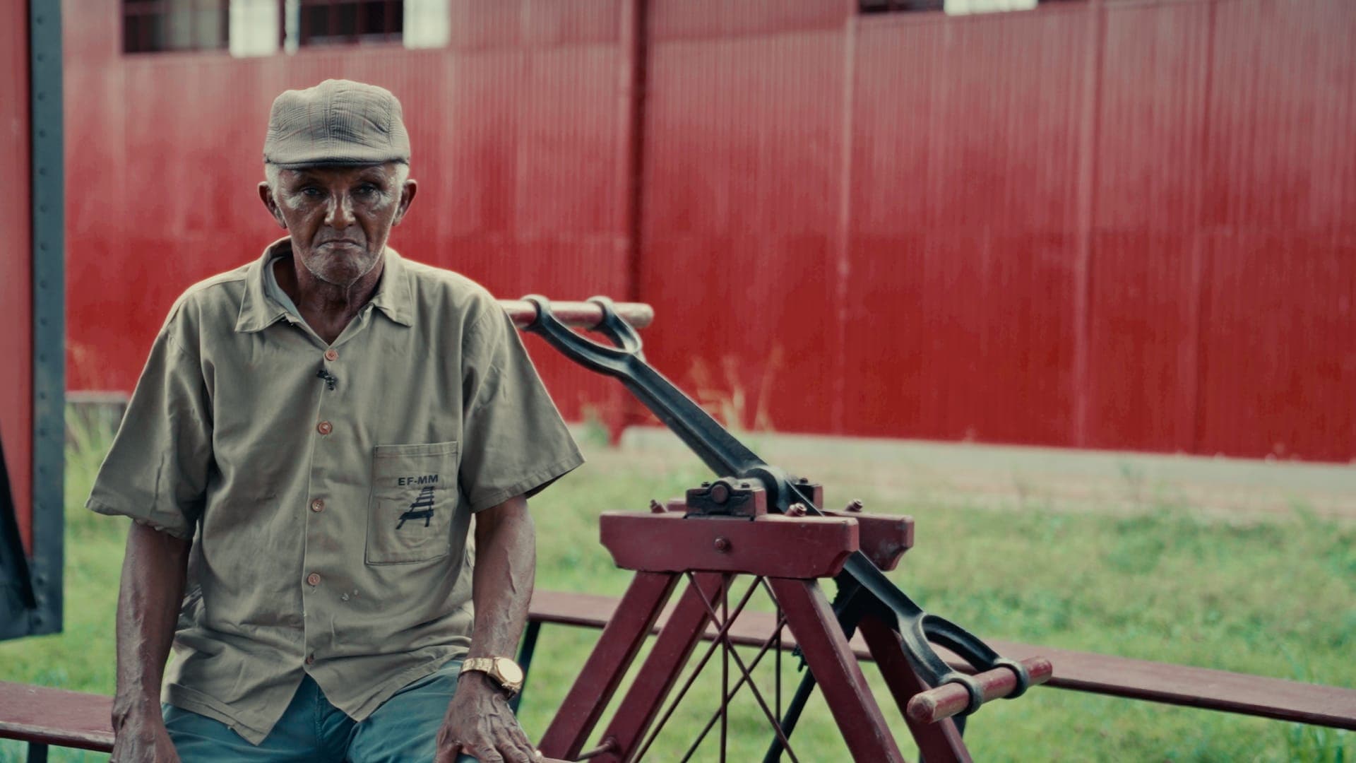 An older man in a cap and khaki shirt beside a red railway handcar, in front of a red shed.