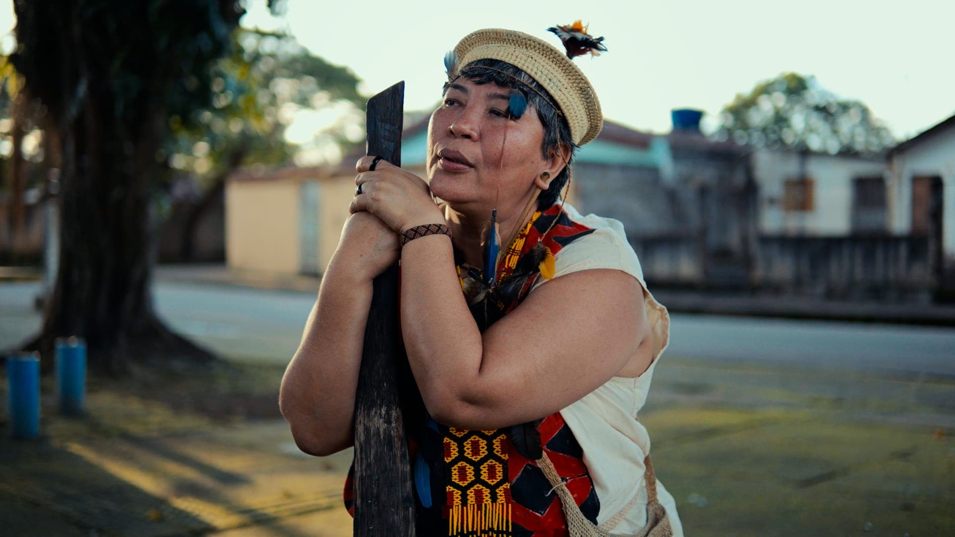 An Indigenous woman in a feather headdress and necklaces holding an object close to her face, on a street.